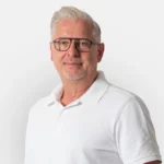 Mann mittleren Alters mit Brille und grauen Haaren, der ein weißes Poloshirt trägt und freundlich in die Kamera lächelt.