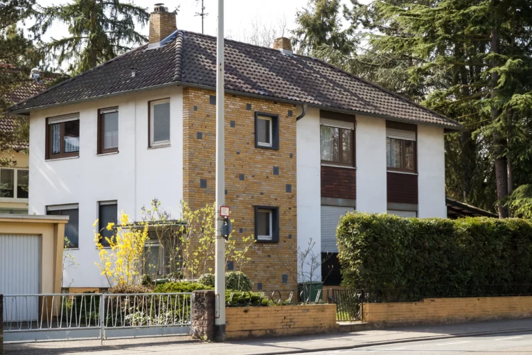 Zweistöckiges Wohnhaus mit hellem Putz und Backsteinfassade, umgeben von Bäumen und grüner Bepflanzung.