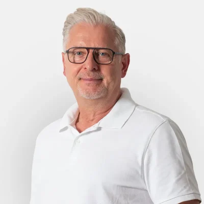 Wilfried-Bosâk Mann mittleren Alters mit Brille und grauen Haaren, der ein weißes Poloshirt trägt und freundlich in die Kamera lächelt.
